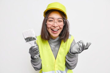 Positive brunette young Asian woman holds paint brush remodels house wears safety equipment and uniform smiles happily isolated over white background. Occupation building and repairing concept