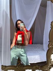 Stylish women at home in bright red sweater and mint green pants take photo selfie in mirror on phone for stories social media, vertical