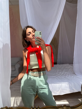 Stylish women at home in bright red sweater and mint green pants take photo selfie in mirror on phone for stories social media, vertical