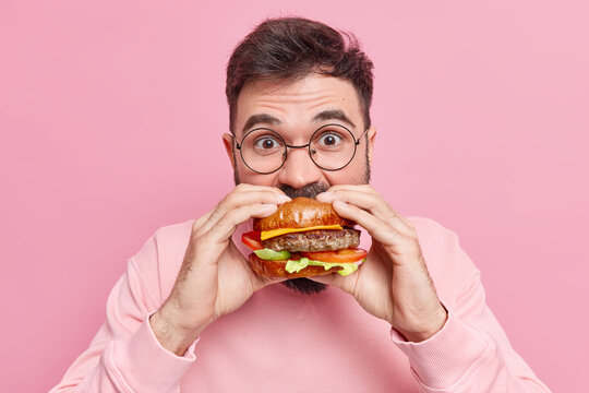 Close up shot of young man eats greedily delicious hamburger feels very hungry consumes fast food wears round spectacles and jumper isolated over pink background. Unhealthy nutrition concept