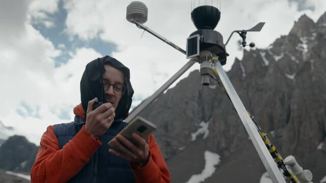 man in glasses in a red hoodie and a blue vest in the mountains checks the functioning of the weather station, a radio message. Mobile weather station. measurement of meteorological conditions