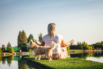 Naklejka premium Plus size woman sitting in lotus position and her cairn terrier dog. Rear view of overweight woman practicing yoga on pier at lake. Leisure activity outdoors
