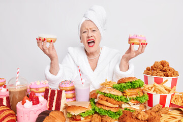 Emotional wrinkled old lady exclaims loudly holds two delicious donuts has bad mood wears white bathrobe and towel over head surrounded by junk food has cheat meal day. Unhealthy eating concept