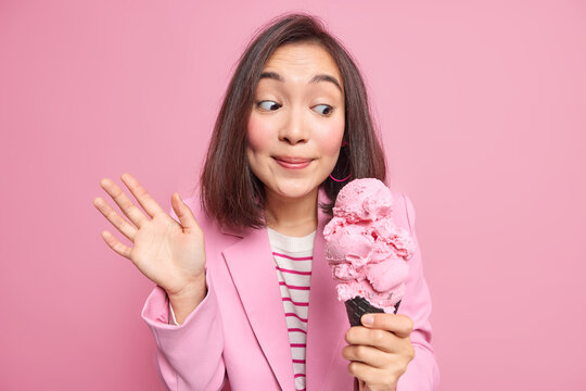 Pretty young Asian female model looks with temptation at tasty ice cream raises palm has sweet tooth cannot imagine her life without delicious desserts wears stylish jacket isolated on pink wall