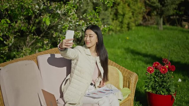 Happy Asian millennial woman taking selfie on smartphone sitting in sunny garden with paperwork and laptop. Portrait of positive slim charming device addicted lady enjoying working from home