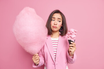 Upset young Asian woman holds delicious ice cream and cotton candy feels temptation to eat sweet food cannot refuse eating tasty dessert dressed in formal jacket isolated over pink background