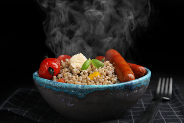 Tasty buckwheat porridge with sausages on table against black background