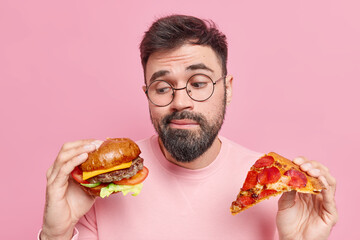 Handsome bearded adult European man feels hesitant whether to eat hamburger or pizza prefers eating...