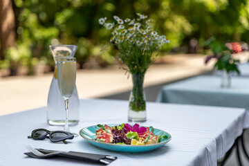 Cafe terrace at sunny summer day. Delicious healthy salad served on outdoor table with white tablecloth, a decanter of cold water with lemon and a vase with a bouquet of chamomile. Selective focus.