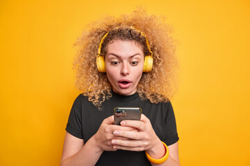 Studio shot of impressed curly haired woman focused at smartphone display feels very surprised reads text message dressed in casual black t shirt isolated over yellow background. Human reactions