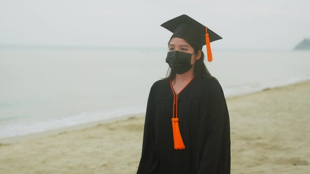 An Asian female graduate who graduated from the university. She walks on a beach with water and waves in the background, wearing a surgical mask to prevent the coronavirus outbreak.