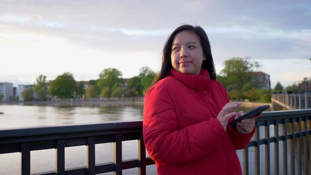 Close up Asian woman wearing red coat standing by the river and looking some good nature in the evening at the park and typing on her phone to have a chat with friend