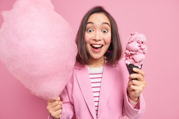 Studio shot of happy surprised young Asian woman with dark hair stares impressed at camera holds cotton candy and tasty appetizing ice cream dressed formally isolated over pink studio background.