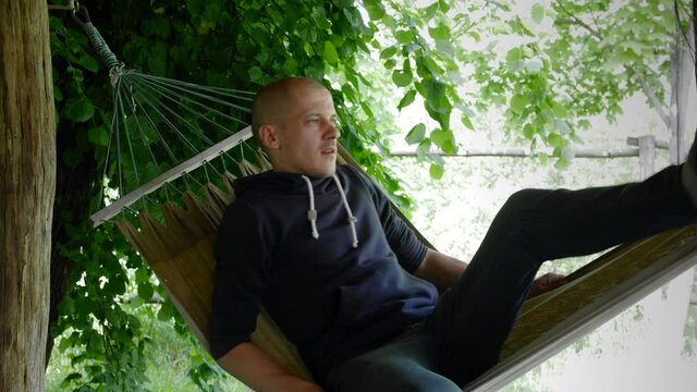 4k stock video footage. A thin, bald Caucasian man walks over to the hammock and lies down. A young man relaxes on a hanging hammock on the terrace of a country house during summer vacation.
