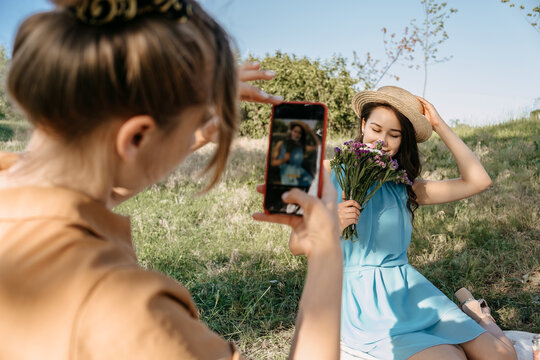 Back view of Young woman holding cell phone and making photo of her girl friend. Two girlfriends take pictures of each other on Smartphone camera on nature background. Summer holidays and technology