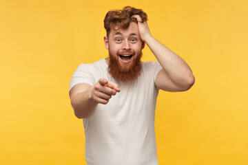 portrait of cheerful positive man with red beard wears blank t-shirt, laughing and point with a finger at the camera. isolated over yellow background