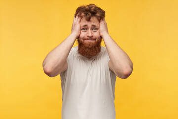 indoor shot of nervous sad man with red hair and beard, wears blank t-shit, holds his head with both hands, pressing his lips and keeps eyes widely opened. isolated over yellow background