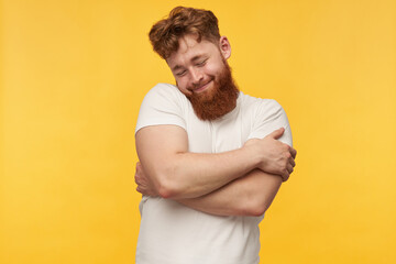 indoor portrait of young bearded guy, wears blank t-shirt, keeps his eyes closed, hug himself and smiling. isolated over yellow background