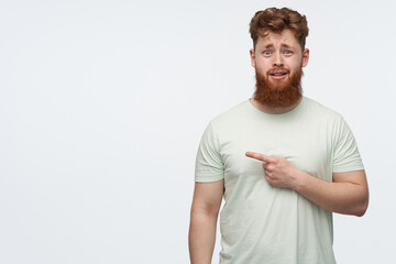 confused guy with red hair and beard wears blank t-shirt, watching into camera and pointing to left...
