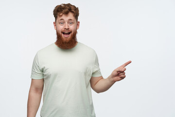 indoor shot of young bearded redhead guy, wears blank t-shirt, pointing with a finger at right side copy space, smiling and has amazed facial expression. isolated over white background.