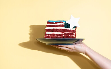 Female hand holding a plate with a piece of cake in the form of the USA flag on a yellow...
