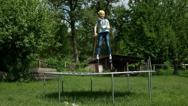 4K colorful spring or summer video footage. A little girl, scholl girl with a broken arm in a plaster cast jumps merrily on a trampoline. Child having fun while on holiday in a country house