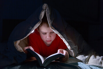 Kid reading book under the blanket in the bed at night. 
