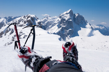 resting on top, feet in crampons and mountaineering ice ax protrude from the snow sparkling in the sun against the backdrop of snowy alps