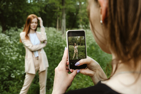 Back view of Young woman holding cell phone and making photo of her girl friend. Two girlfriends take pictures of each other on Smartphone camera on nature background. Summer holidays and technology
