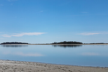 Sandy beach by still water, distant islands, blue sky reflected in the water, horizontal aspect