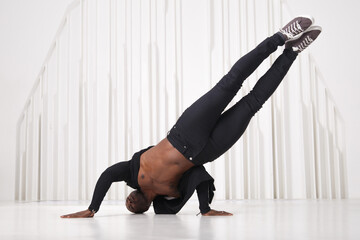 Elegant black man dancer in black clothes does a handstand in a bright room.