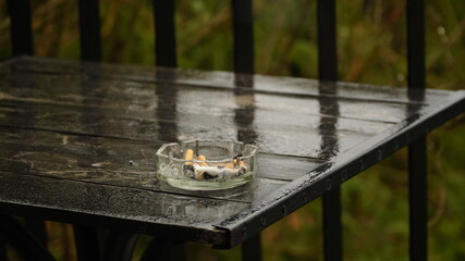 Glass ashtray. Wet ashtray with extinguished cigarettes on the table after the rain. Bad habits concept.