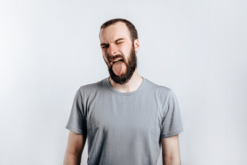 Handsome young brunette man frowning and closing eyes and sticking out tongue, angry on white background with space for advertising mock up