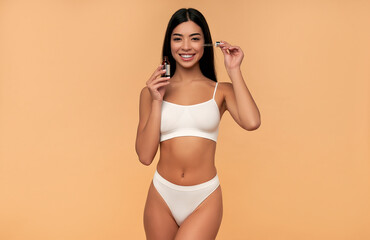 Naklejka premium Young Asian woman with clean radiant skin in white lingerie uses a moisturizing serum on a beige background. Spa care, facial skin care, beauty cosmetology.