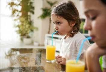 Beautiful baby girl drinking orange juice. Blurry boy on the foreground
