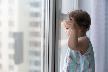 Small pensive girl against closed window looking out. Quarantine and self-isolation concept. Copy space