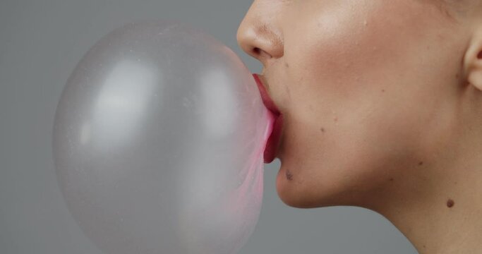 Young woman blowing a bubble with gum on grey background, closeup