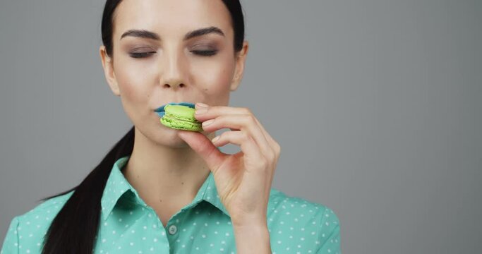 Beautiful young woman with unusual makeup eating sweet macaron on grey background