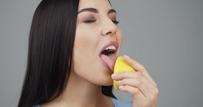 Beautiful young woman licking half of soar lemon on grey background