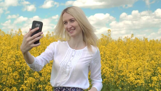 Portrait of a happy woman taking a selfie and raising her hand in a field of yellow rapeseed flowers. She smiles and takes photos on her smartphone.