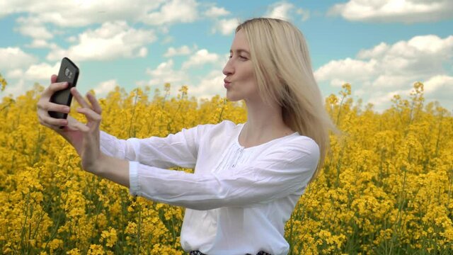 a young happy blonde in a white blouse and skirt, using a mobile phone for a selfie on a yellow rapeseed flower field. She is happy and enjoys life. He smiles.