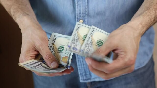 Unrecognizable caucasian man in jeans and a denim shirt stands, counting out dollar bills, puts in money clip. Only hands visible. Paper money, cash. Side view. Finance, budget concept. Horizontal
