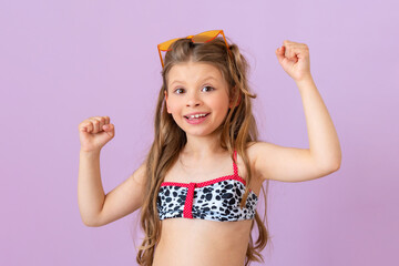 A beautiful little girl in a swimsuit and sunglasses shows her fists.