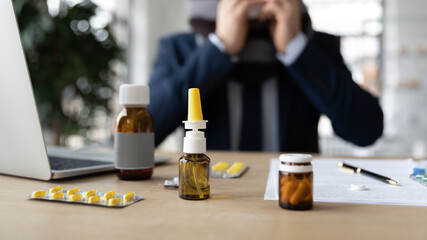Workplace desk of sick businessman, office employee suffering from flu. Table top with laptop, glass bottles of drugs, nasal drops, blisters of pills. Influenza treatment, remedy, therapy concept