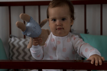 Cute cheerful little chubby baby girl sitting in grey baby crib after waking up from sleep in white...