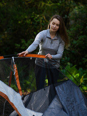 In the photo, a young woman with long blond hair is traveling, resting in the forest, laying out a tent. In the background is a photo of a dense green coniferous forest.