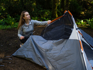 In the photo we see morning in the forest. Camping. Young woman folds up the tent. The woman looks up. In the background of the photo we see a dense forest.