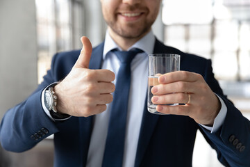 Happy businessman recommending drinking fresh pure water for refreshment, hydration balance and wellbeing, holding glass and making thumb up like gesture. Water purification, healthcare concept