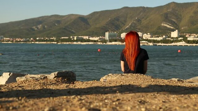 a girl with red hair sits on the beach, her back to the camera