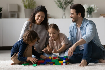 Overjoyed multiethnic parents sit on floor play toys with little biracial kids together. Happy multiracial young family with small ethnic children have fun engaged in playful game activity at home.